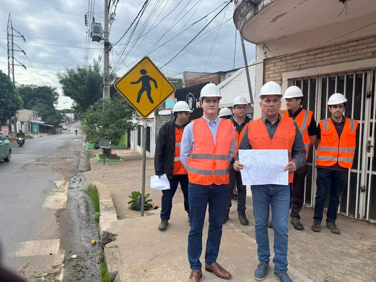 Dos hombres discuten planos, rodeados de otros cinco en chalecos naranjas y cascos de seguridad, en una calle con cartel de tráfico.