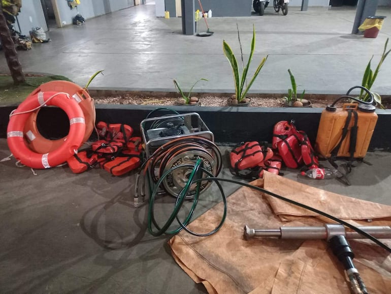 Varios chalecos salvavidas rojos y un salvavidas naranja organizados en un área de almacenamiento iluminada con mangueras y equipo.