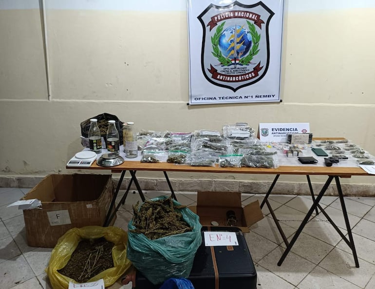 Mesa con paquetes y bolsas de marihuana, balanza y teléfono móvil en oficina de Policía Nacional, sin personas visibles.