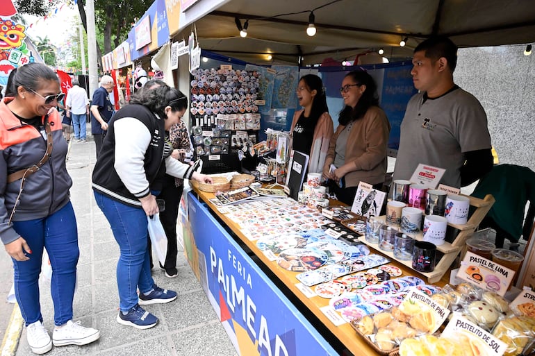 Mujer con gafas de sol sonríe mientras sostiene una bolsa, rodeada de productos coloridos en el mercado festivo.