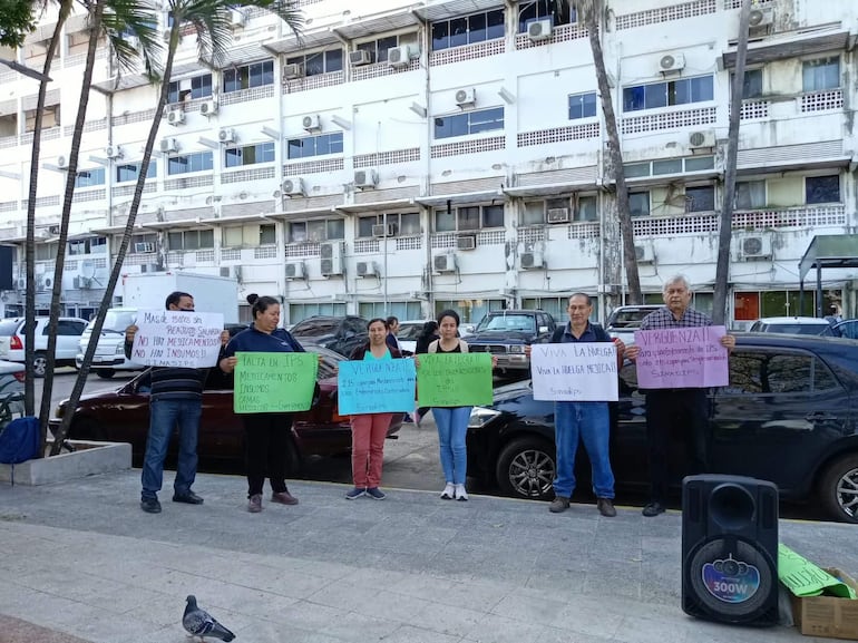 Seis manifestantes sostienen carteles sobre falta de insumos y salario, en una protesta consciente frente a un edificio urbano.