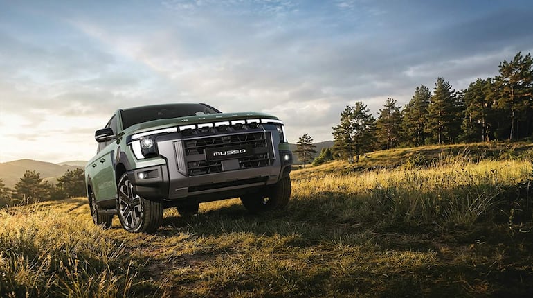SUV MUSSO verde en primer plano, con colinas y árboles de fondo bajo luz natural.
