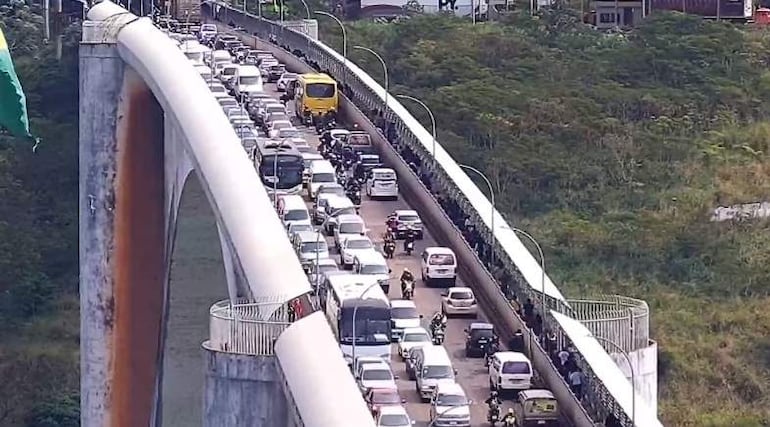 Tráfico detenido en el puente, con motocicletas en el lateral y vegetación al fondo.