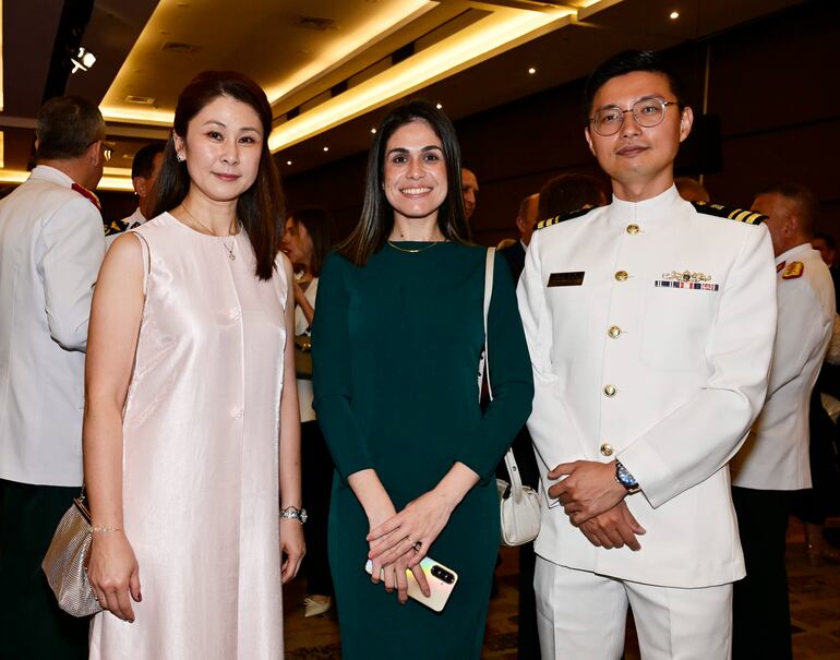 Dos mujeres sonrientes, una con vestido rosa claro y otra con vestido verde oscuro, junto a un hombre en uniforme blanco en evento formal.