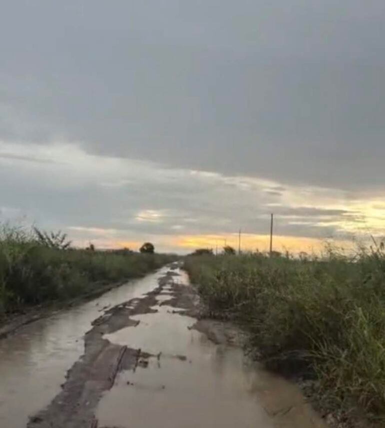 Camino de tierra en mal estado con charcos, rodeado de vegetación y postes eléctricos. Cielo nublado al atardecer.