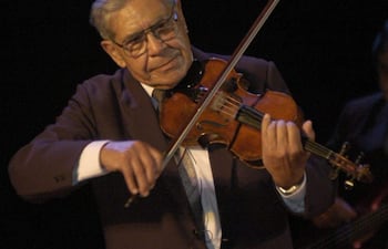 Hombre mayor con traje oscuro y gafas, tocando el violín con expresión concentrada en un ambiente oscuro.
