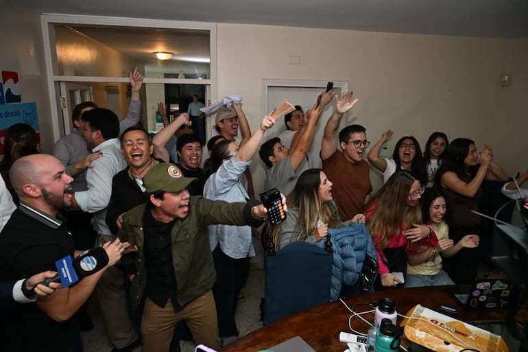 Grupo de personas con gestos de alegría en reunión cerrada, un hombre con micrófono y otro con gorra verde celebran la victoria.