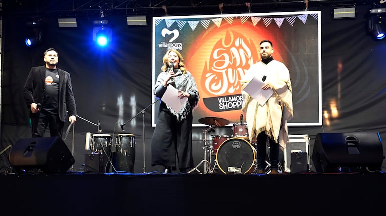 Presentadores en escenario: hombre con chaleco negro, mujer en vestido negro y poncho gris, hombre con poncho claro, instrumentos musicales al fondo.