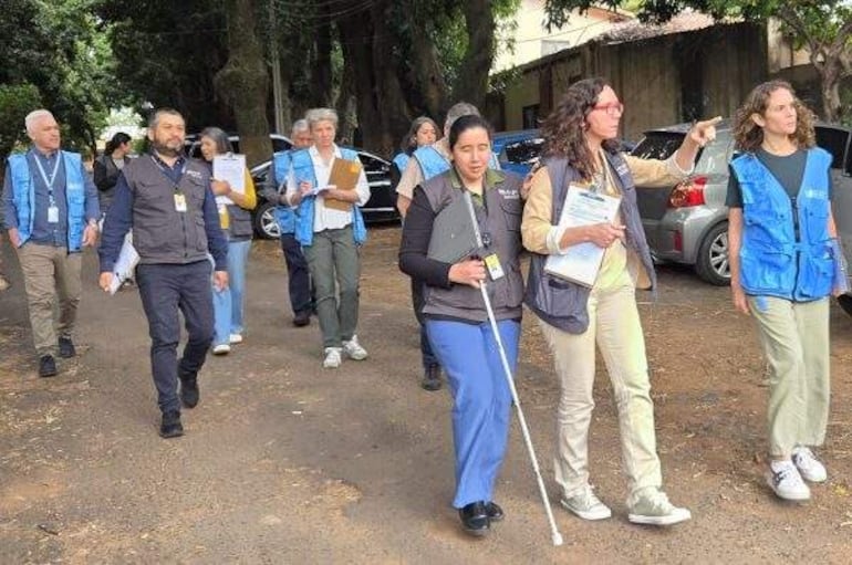Grupo de diez personas, en su mayoría con chalecos, camina por un terreno de tierra, algunas portando documentos. Árboles al fondo.