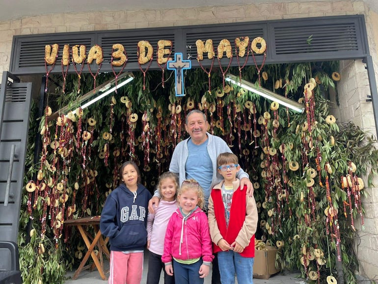 Hombre de camiseta azul y abrigo claro sonríe rodeado de cuatro niños en un evento festivo con decoraciones verdes.
