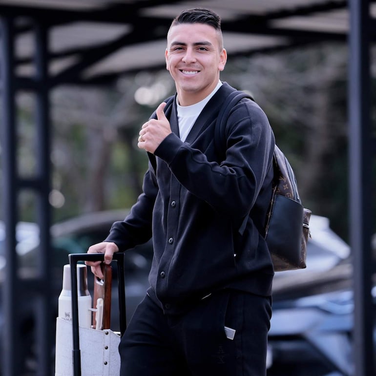 Braian Ojeda sonríe con pulgar hacia arriba, vestido con suéter negro y pantalones deportivos, sosteniendo un equipaje sobre ruedas.