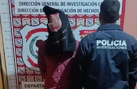 Hombre encapuchado entre policías en sede de investigación, uno lleva chaqueta marcada con 'POLICÍA INVESTIGACIONES'.