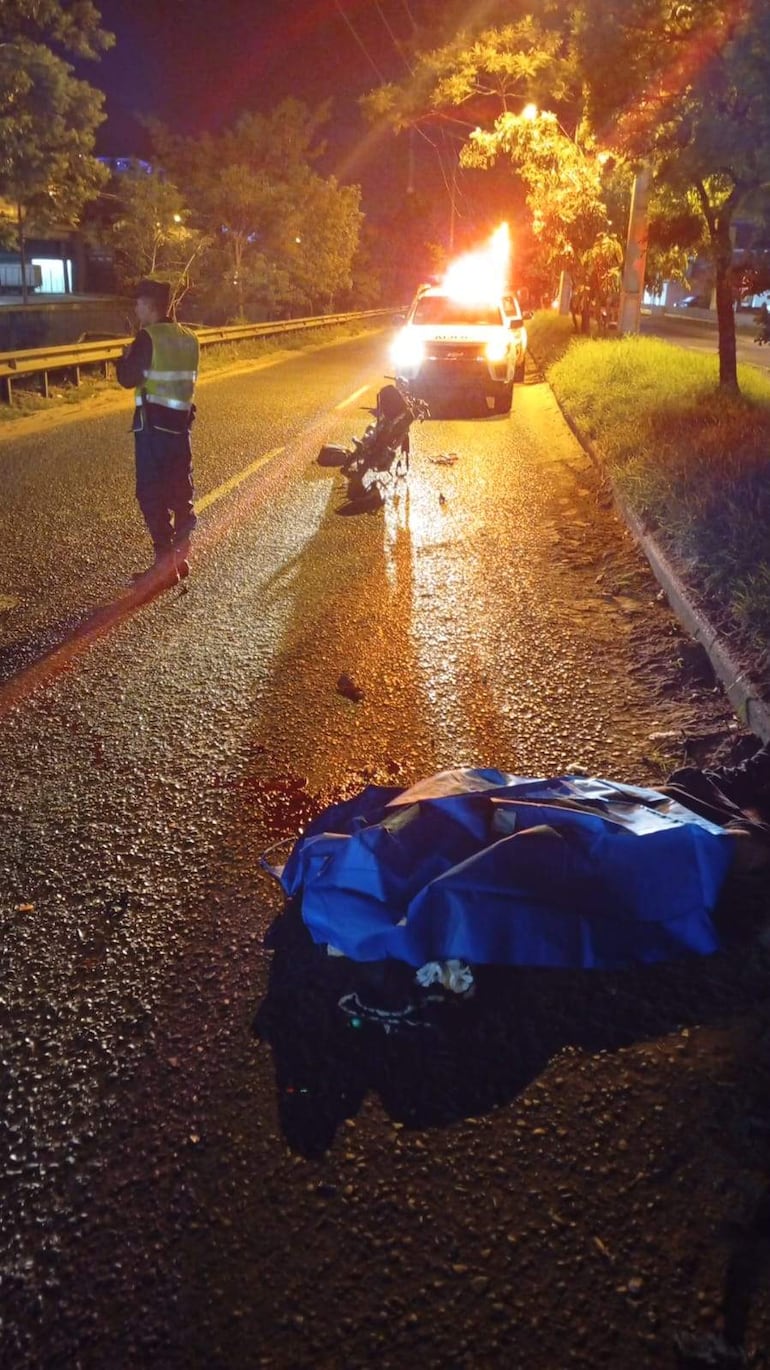 Un oficial de seguridad en chaleco reflectante junto a una motocicleta caída, con un objeto cubierto y un vehículo policial iluminado al fondo.