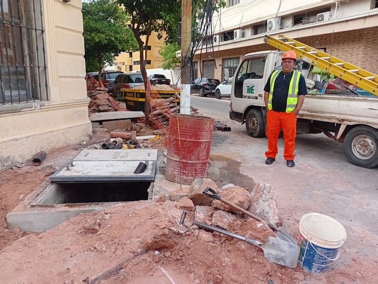 Hombre con casco anaranjado y chaleco reflectante en obra con tierra removida y materiales de construcción.