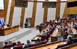 Óscar Lovera en podio junto a mujer de abrigo amarillo y otros oradores, con audiencia atenta en ambiente gubernamental.