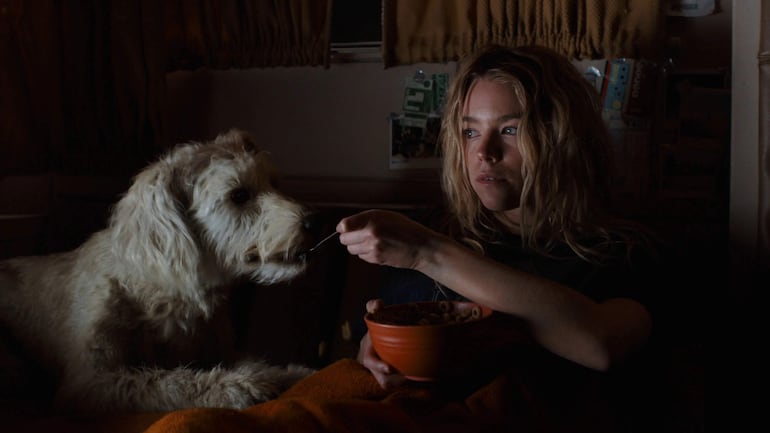Milly Alcock, con cabello largo y desordenado, sentada con un recipiente de cerámica en mano, mientras su perro espera comida.