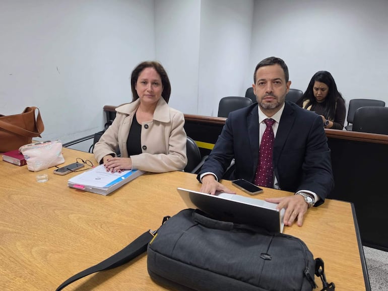 Francisco Cabrera con traje negro, corbata de puntos y expresión seria, junto a mujer en abrigo beige, en mesa con documentos y celular.