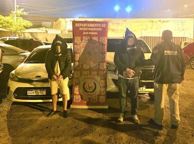 Tres hombres, con capuchas, son supervisados por un policía en chaqueta, bajo un cartel del departamento de investigaciones, en un entorno nocturno.