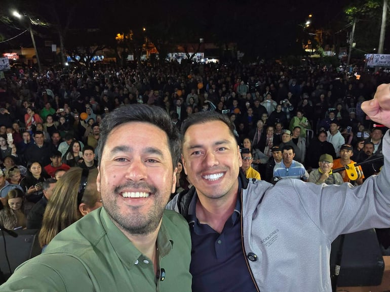 Miguel Prieto con chaqueta gris y Eduardo Desjesus en camisa verde sonriendo para una selfie, rodeados de una multitud animada.