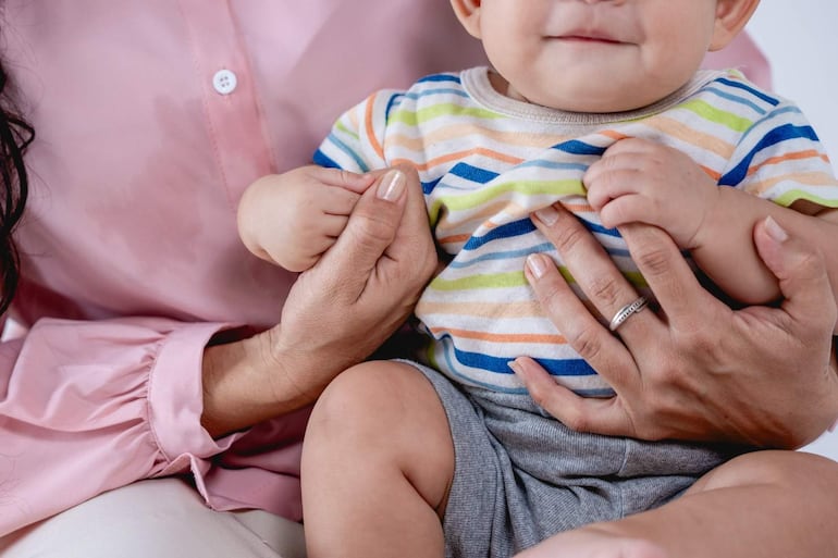 Mujer con camisa rosa sostiene a un niño en camiseta a rayas mientras ambos miran hacia la cámara en un ambiente neutro.