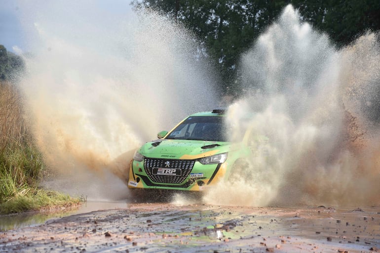 Coche de rally verde con detalles amarillos salpicando barro mientras avanza rápido en pista natural.