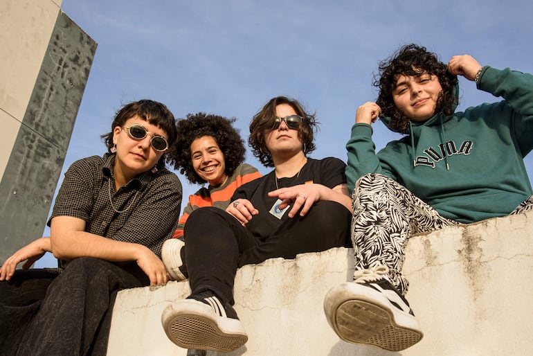 Cuatro mujeres sentadas, una con gafas de sol y camiseta de rayas, otra sonriente con cabello rizado, bajo un cielo despejado.