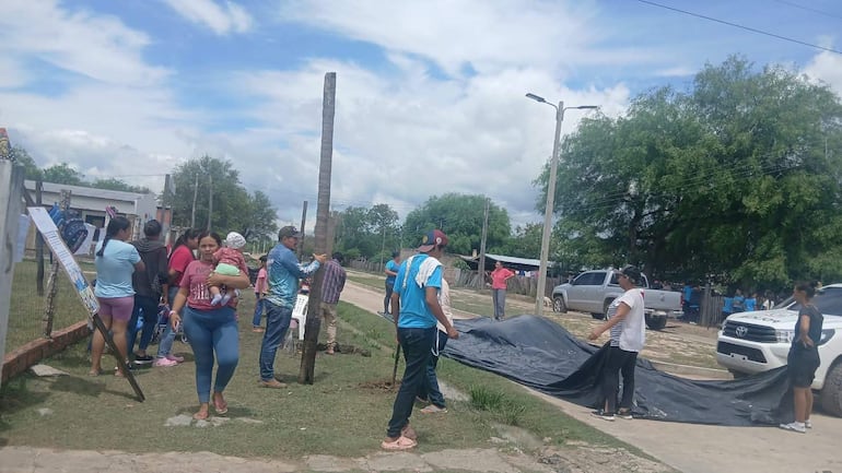 Varias mujeres, niños y un hombre con sombrero y camisa azul interactúan en un ambiente tranquilo con un vehículo policial al fondo.