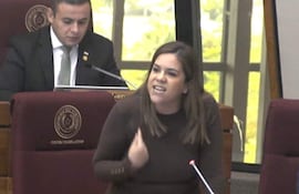 Johanna Ortega gesticula con expresión enfadada en un entorno legislativo, un hombre la observa en segundo plano.