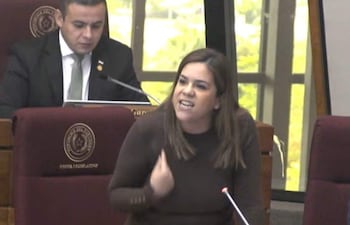 Johanna Ortega gesticula con expresión enfadada en un entorno legislativo, un hombre la observa en segundo plano.