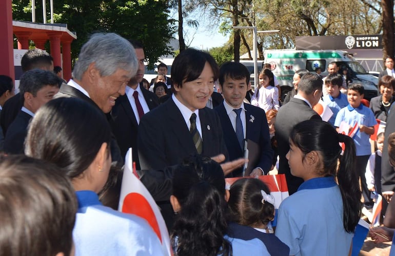 El príncipe Akishino sonriente conversa con niños que sostienen banderas en un evento festivo al aire libre.
