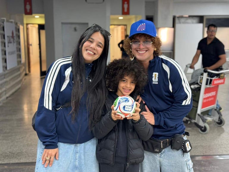 Jennifer Hicks y Miguel Narváez sonríen, vestidos con chaquetas deportivas, mientras un niño sostiene un balón de fútbol en un ambiente moderno.