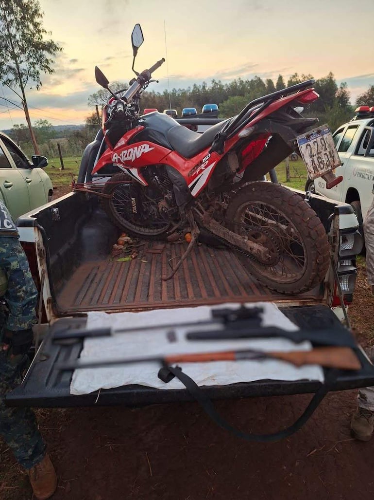 Camioneta en área rural con motocicleta roja y arma sobre tela blanca, sin personas visibles, sugiere operación de seguridad.