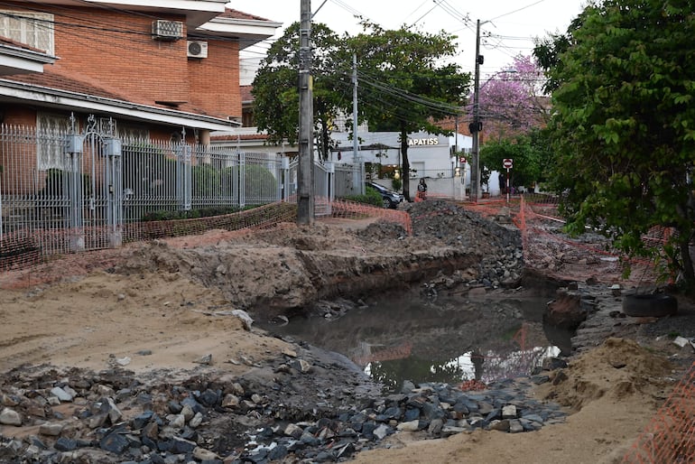 Calle con obra en construcción, cercada con malla naranja y tierra excavada, sin personas visibles.