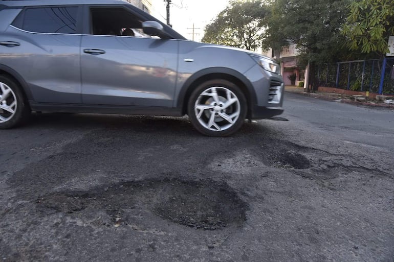 Automóvil gris en movimiento sobre bache en calle, con edificios y árboles de fondo.