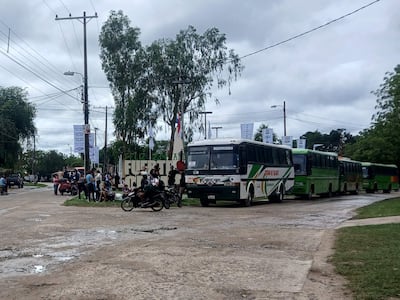 Grupo de personas esperando transporte en Fuerte Olimpo, con clima nublado y autobuses estacionados visibles.