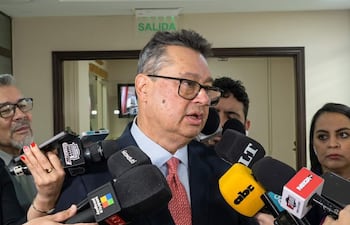Gustavo Leite con gafas y traje oscuro rodeado por periodistas en un entorno cerrado, visiblemente serio y concentrado.