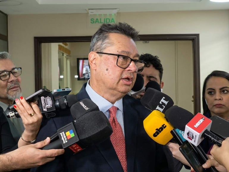 Gustavo Leite con gafas y traje oscuro rodeado por periodistas en un entorno cerrado, visiblemente serio y concentrado.