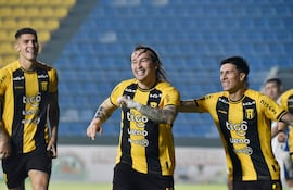 Tres jugadores del Club Olimpia sonríen y celebran, vestidos con camisetas amarillas y negras, en un estadio con gradas ocupadas.