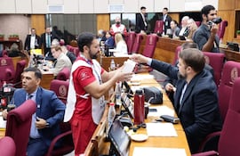 Pedro Alejandro Díaz Verón en camiseta roja entrega un objeto a Orlando Penner Durksen con traje oscuro, en un aula legislativa con sillas burdeos.