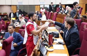 Pedro Alejandro Díaz Verón en camiseta roja entrega un objeto a Orlando Penner Durksen con traje oscuro, en un aula legislativa con sillas burdeos.