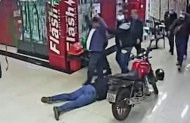 Hombre tendido en el suelo, vestido de azul, rodeado por cuatro hombres en un ambiente de tensión en un comercio.