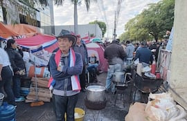 Hombre con sombrero y brazos cruzados observa protestas de adultos mayores en ambiente urbano, rodeado de banderas paraguayas.