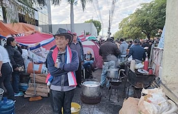 Hombre con sombrero y brazos cruzados observa protestas de adultos mayores en ambiente urbano, rodeado de banderas paraguayas.