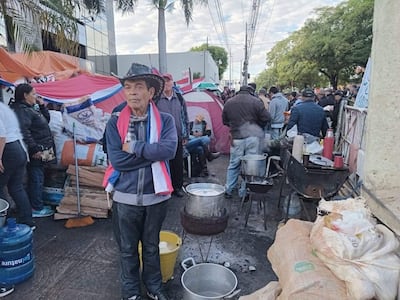 Hombre con sombrero y brazos cruzados observa protestas de adultos mayores en ambiente urbano, rodeado de banderas paraguayas.