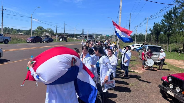 Grupo de bioquímicos con batas blancas y pancartas en protesta en camino rural, algunos sostienen una gran bandera paraguaya.