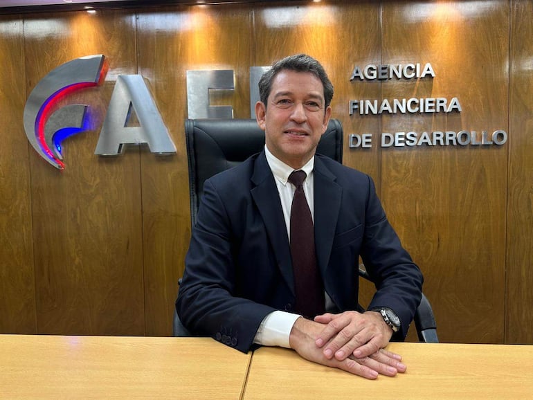 Hombre con traje oscuro y corbata marrón sentado en oficina con logo de la AFD en el fondo, mostrando postura relajada.