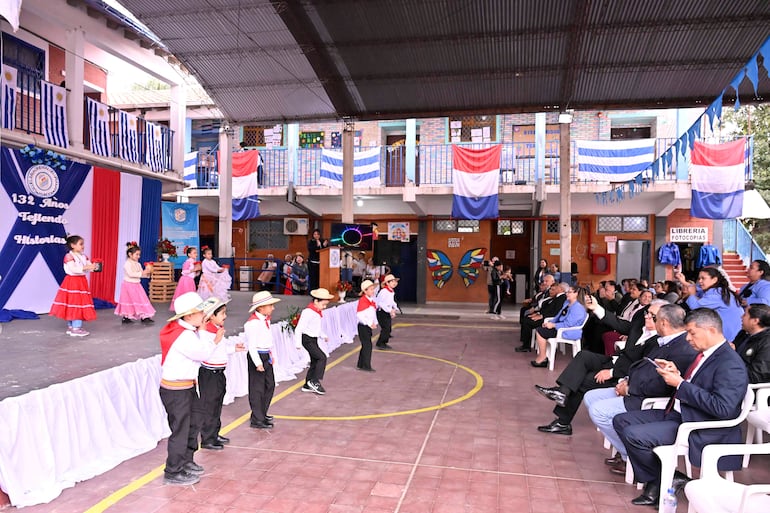 Niños en trajes típicos bailan en un escenario decorado, mientras adultos observan con atención.