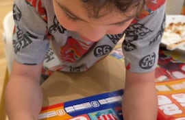 Niño pequeño de cabello rizado, camiseta con personajes, sostiene tarjetas en una mesa en un ambiente interior.