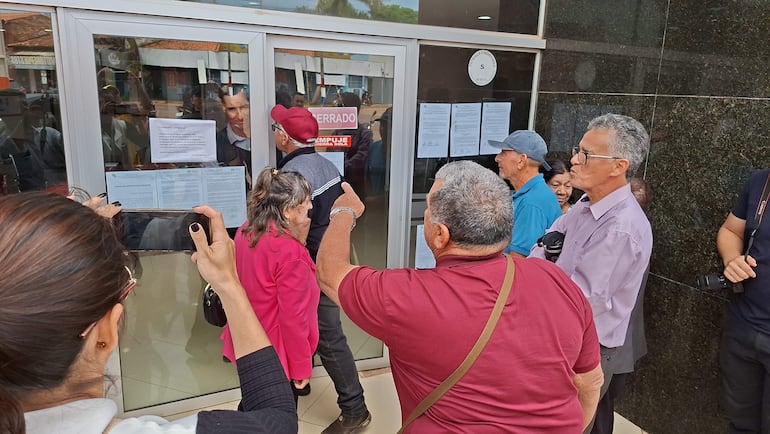 Ahorristas, incluida una mujer con cabello recogido y un hombre de gorra azul, se aglomeran frente a la puerta cerrada de la cooperativa.