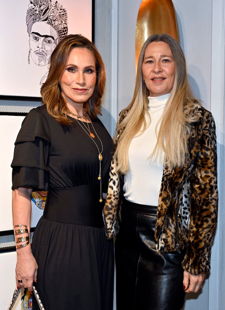 Dos mujeres posan en un entorno interior, una en vestido negro y otra en blusa blanca con abrigo de piel de leopardo.